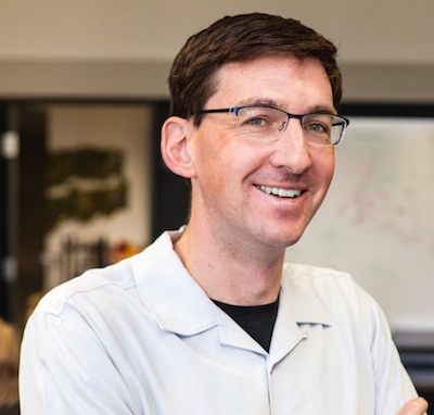 A headshot of Chris Potts wearing a subtly cool hawaiian shirt, with a blurry whiteboard in the background behind him