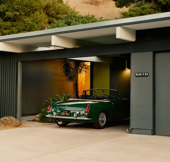 Фото одноэтажного дома в пустыне с крытой парковкой, на которой стоит винтажный спортивный кабриолет.
