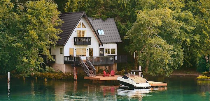 A photo shows a boat, covered and docked in front of a 2-storey home on the edge of a lake.