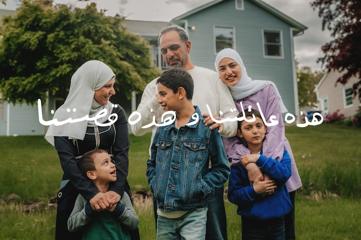 Μια οικογένεια έξι Σύριων προσφύγων στέκεται μαζί, χαμογελώντας στην πίσω αυλή ενός μπλε σπιτιού.