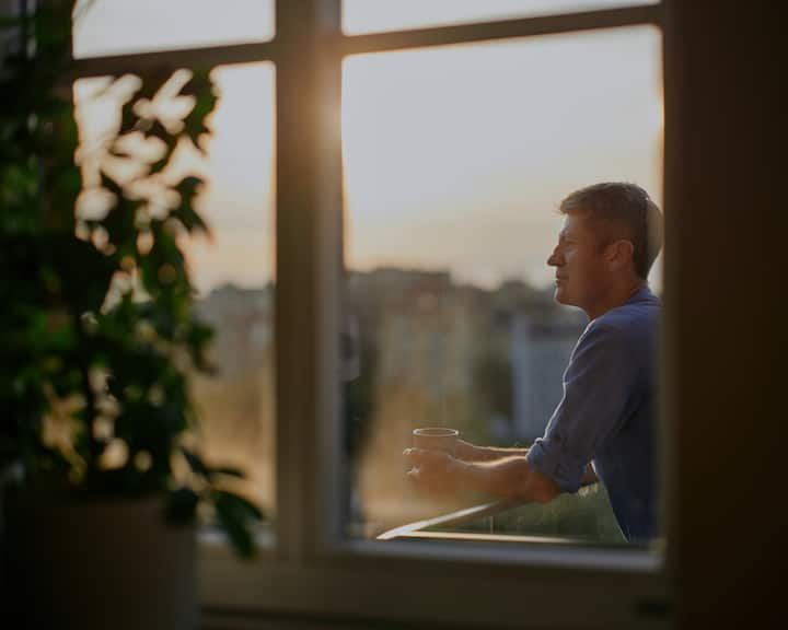 Një burrë me këmishë blu mban në dorë një filxhan kafeje ndërsa qëndron në ballkonin e tij me vështrimin drejt horizontit.