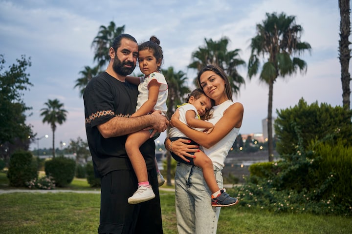 Un tată și o mamă zâmbesc senini, ținându-și în brațe cei doi copii într-un parc cu palmieri.
