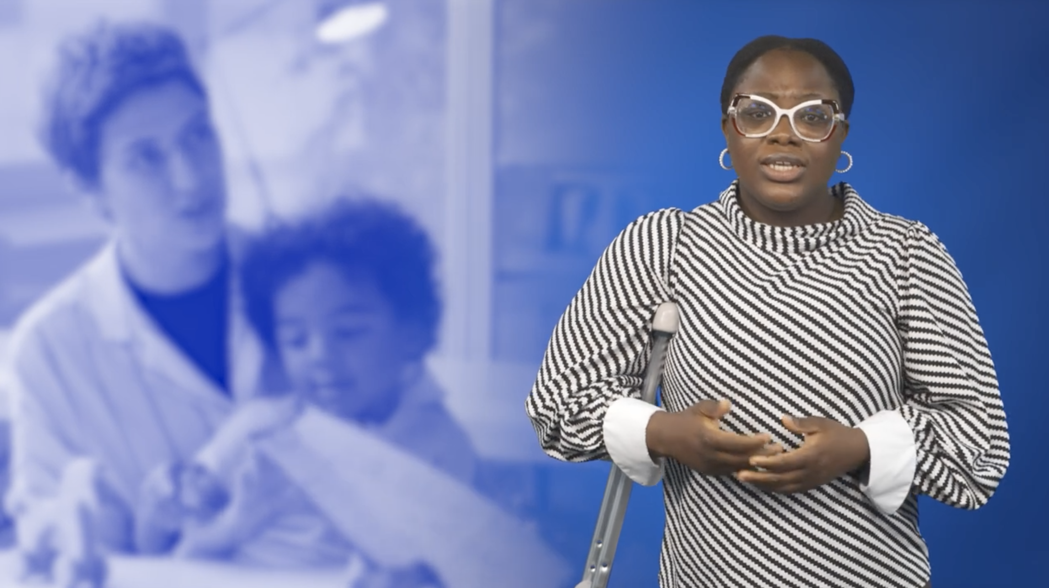 Woman speaks to a camera. A care worker and a child show in the background.