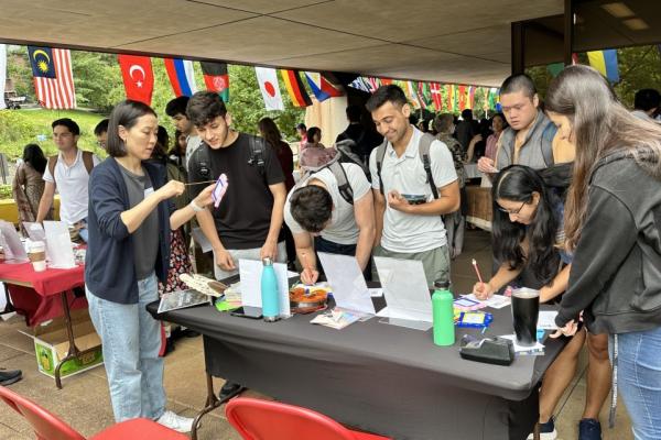 Several people cluster around a table with international flags in the background