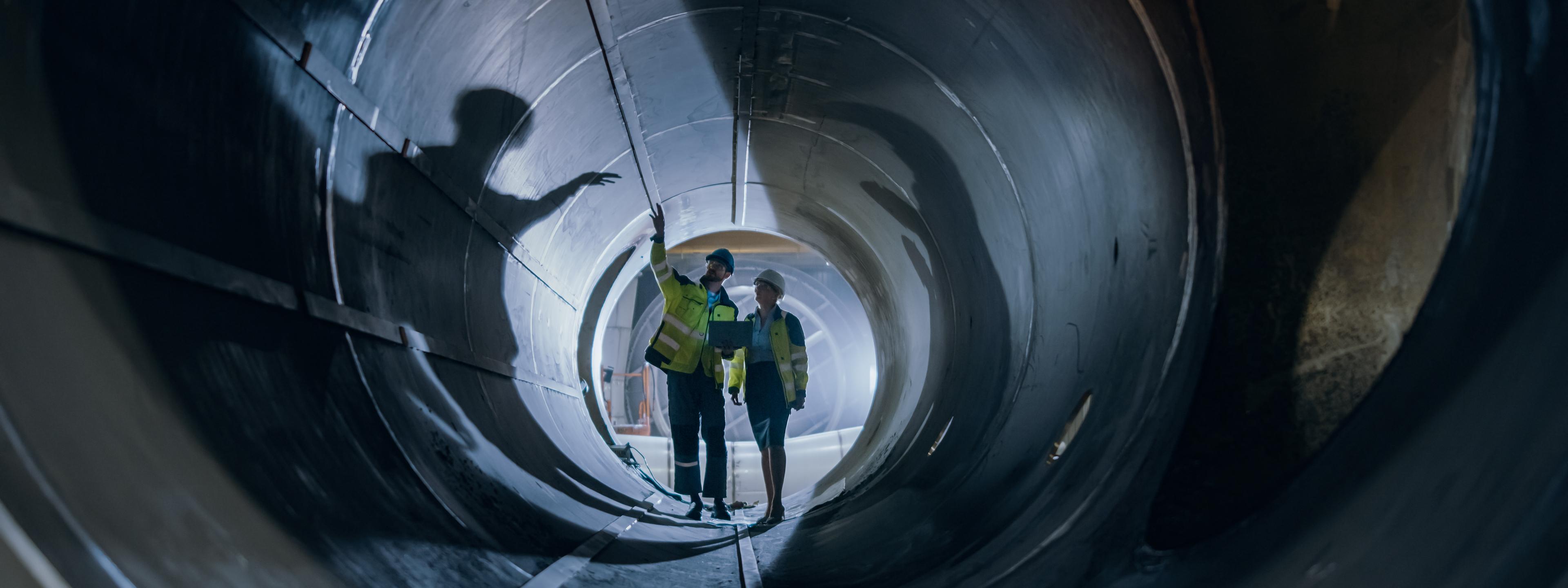 Two Heavy Industry Engineers Walking Inside Pipe, Use Laptop, Have Discussion, Checking Welding. Construction of the Oil, Natural Gas and fuels Transport Pipeline. Industrial Manufacturing Factory