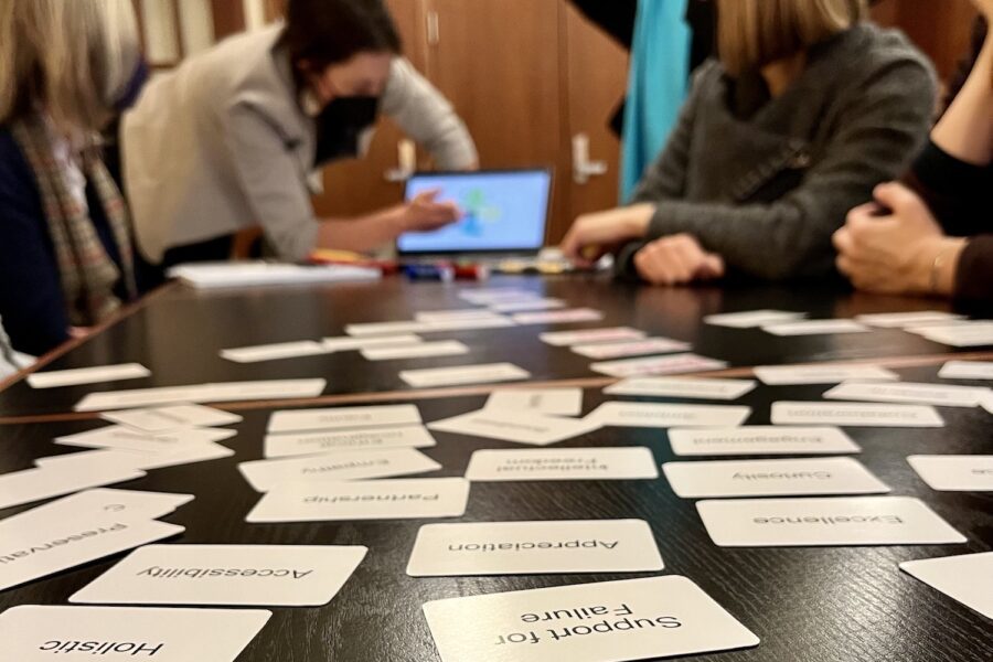 In the foreground in focus are white values cards with black letters laying on a dark wooden table with words like "Support for Failure," "Holistic," "Accessibility,"Excellence," "Appreciation," written on them. We read the cards upside-down as we look down the table, our perspective at table-level looking toward a woman in a black mask leaning over a computer pointing to something as participants around the table look toward the laptop on the table. The laptop and the people are out of focus at the depth of field is short and the focus is on the cards nearest us on the table.