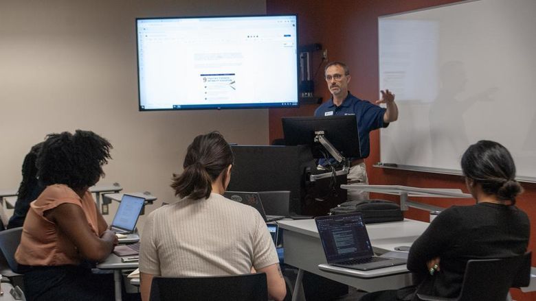 Four people sit in a semicircle in a classroom listening to speaker and watching a screen