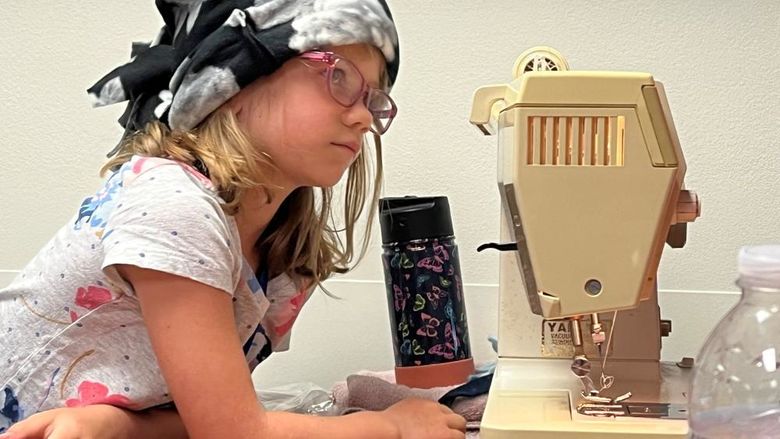 A girl in a fleece winter hat leans over a sewing machine.