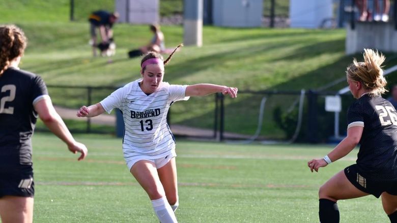 A Penn State Behrend soccer player dribbles between two defenders.