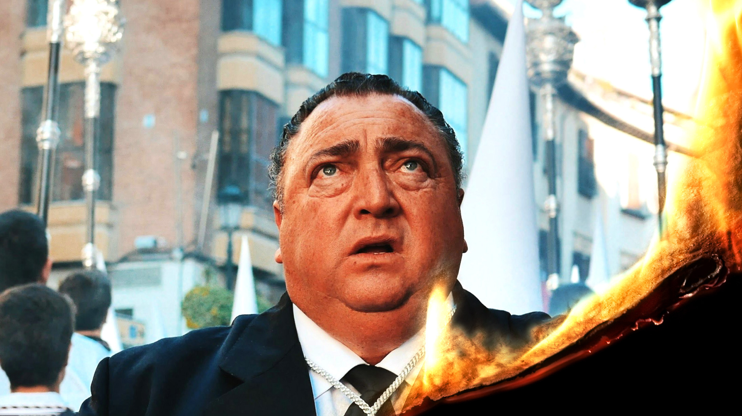 A man in a suit looks upward with a surprised expression. There is a fiery effect in the foreground. Buildings and people are visible in the background.