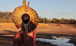 Fotografia de um homem indígena de costas, observando o horizonte. Ele usa um cocar colorido e diversos adereços tradicionais. Ao seu lado, há um pequeno lago cercado por um campo aberto com diversas vegetações. O céu acima está claro e azul.