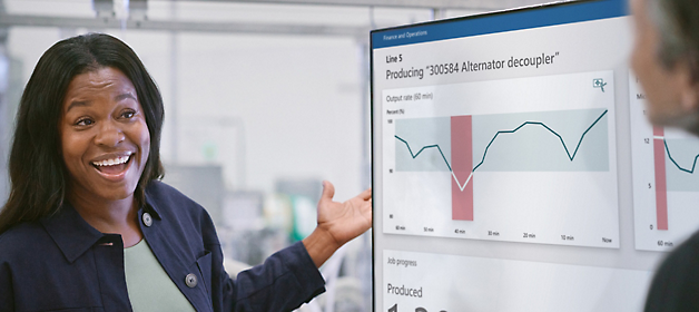 A woman presenting in a meeting with a man and digital screens displaying data in the background. A smiling woman gestures towards a screen displaying production data graphs 