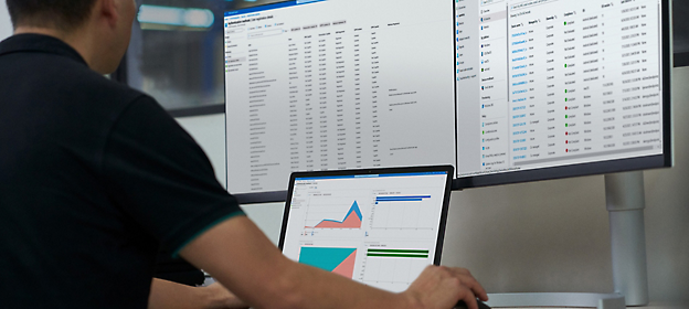 A person working on a laptop with data analysis graphs on the screen, in front of a larger monitor 