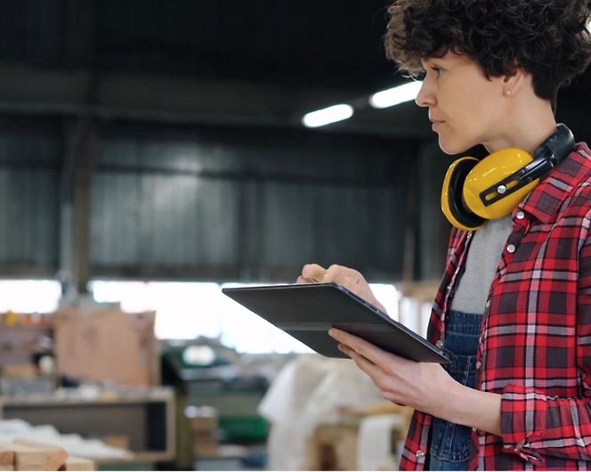 Person in checkered shirt holding a tablet and wearing yellow ear protection around the neck