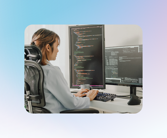 A person in a grey shirt, seated on an office chair, types code on a keyboard in front of two large computer