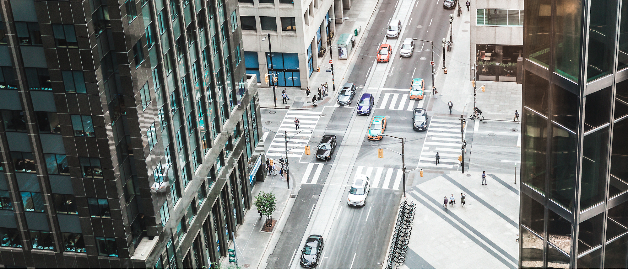 Aerial view of a city street with a few cars and pedestrians at a crosswalk, flanked by tall buildings.