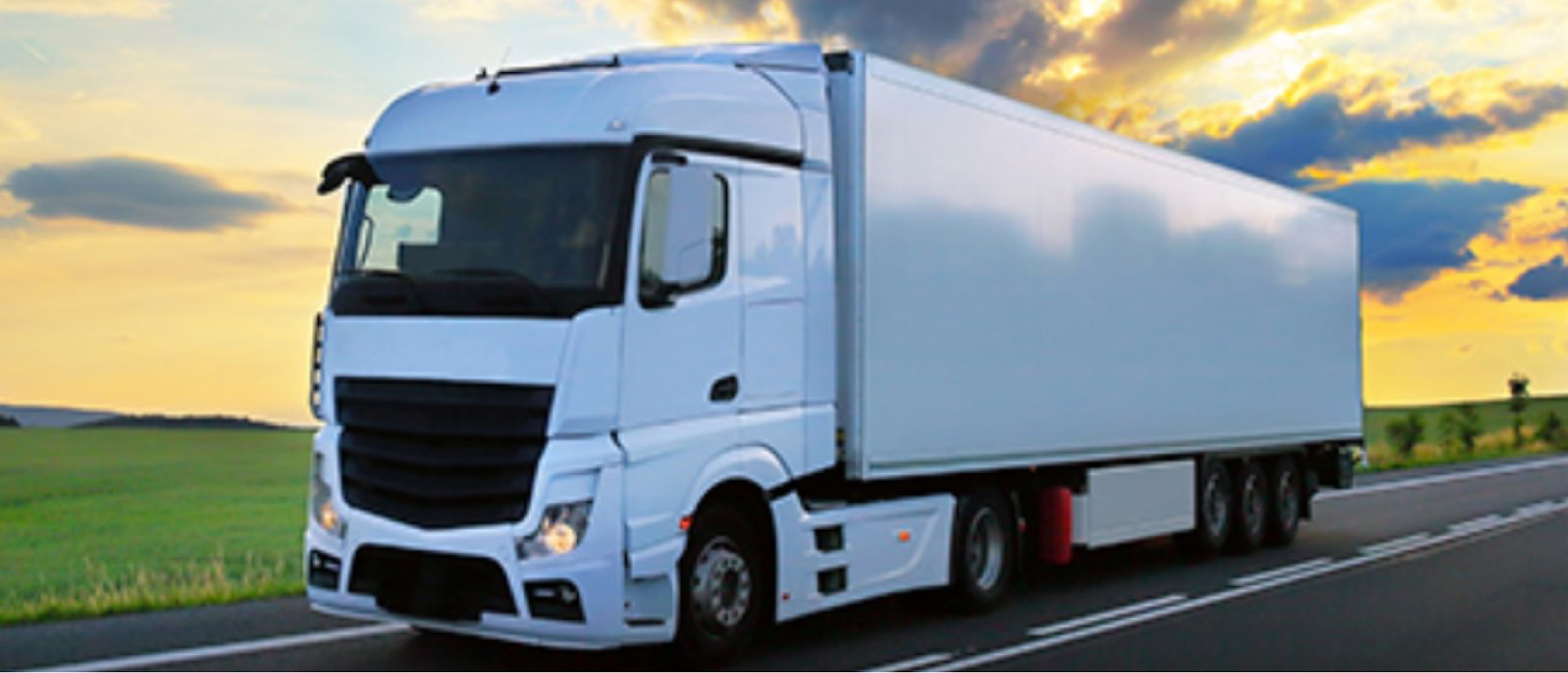 A white semi-truck with a trailer drives down a highway at sunset