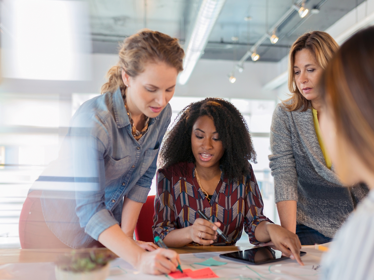 Four women collaborating over documents on a table in a brightly lit office.