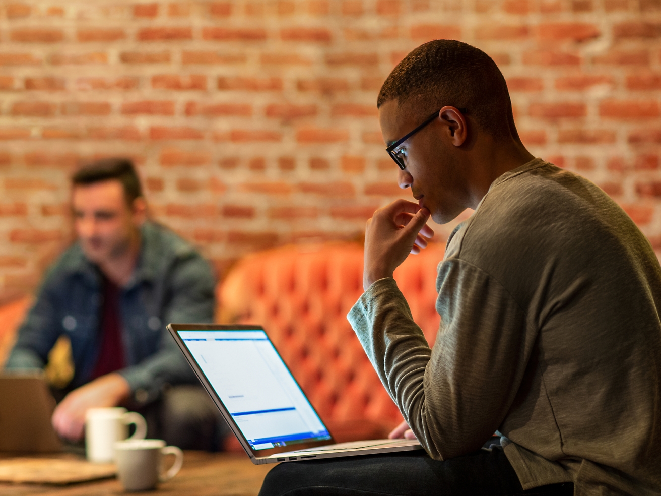 A focused man wearing glasses uses a laptop in a casual office setting with another person in the background.