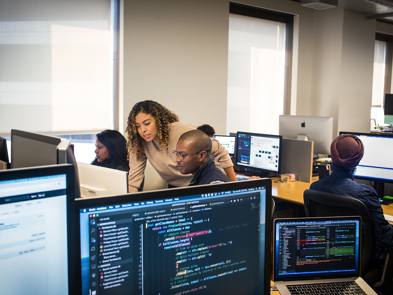 Diverse group of professionals working on computers in an office environment, focusing on screens with various code and data visible.