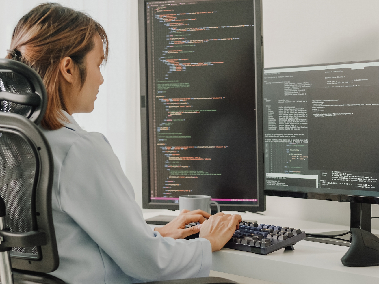 A woman sitting in an office chair, coding on a computer with multiple screen displays showing programming code.