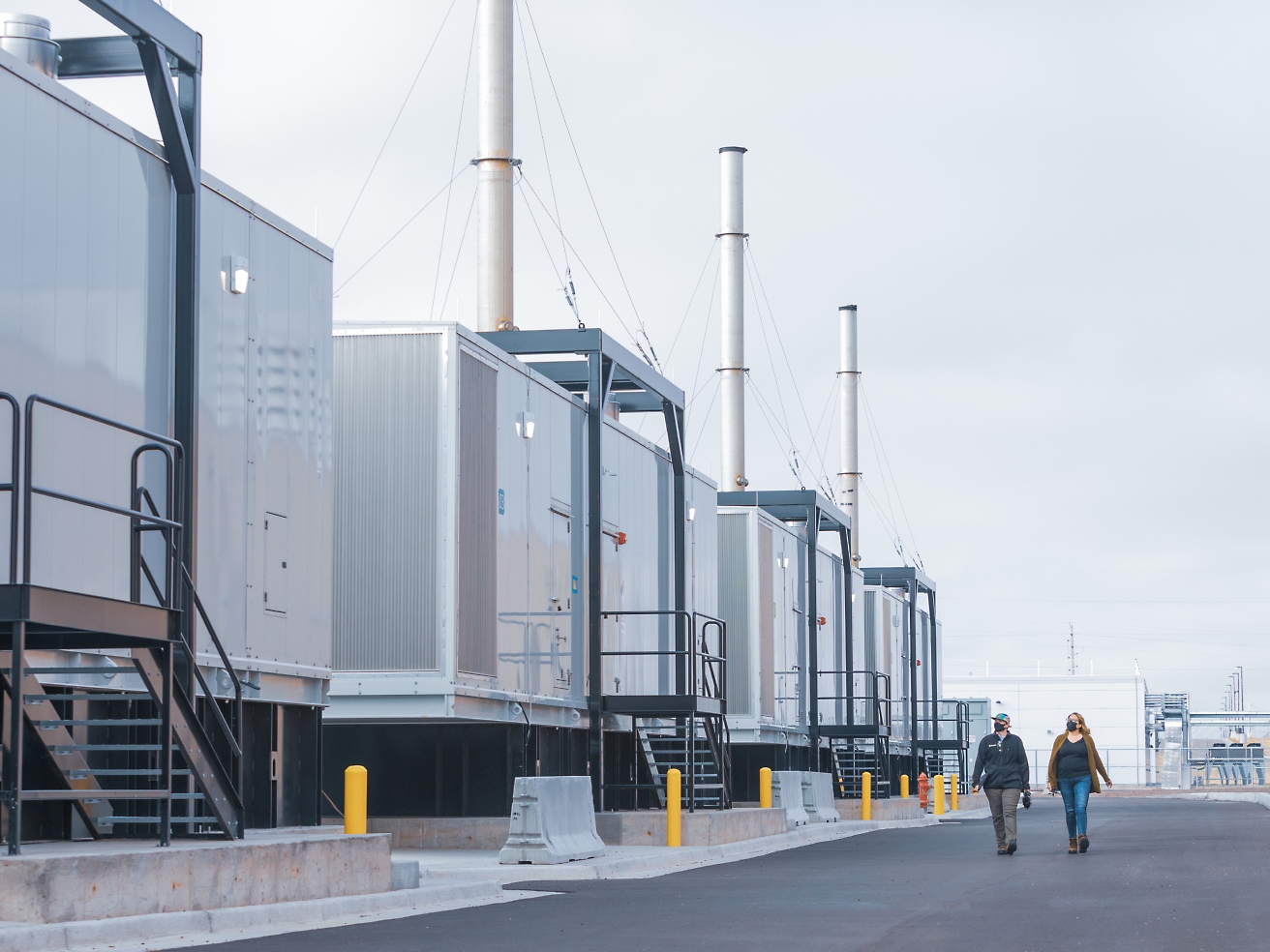 Two people walking by a row of industrial containers with tall chimneys at a facility, under an overcast sky.