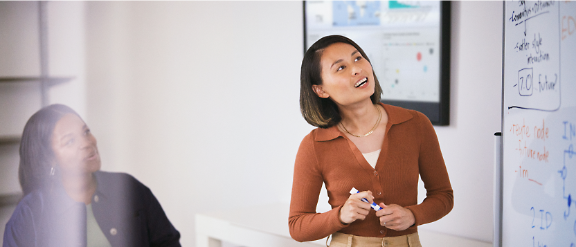 A woman giving a presentation using a whiteboard, with an attentive listener in the background.