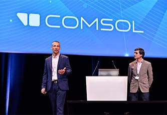 Two speakers presenting onstage, with a podium between them; a slide showing the COMSOL logo is visible behind the speakers.