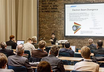 A classroom of people listening to an instructor who is sitting in front of a slide that shows information about electron beam divergence simulation.