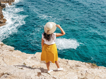 Young woman in yellow skirt standing on edge of cliff above sea.