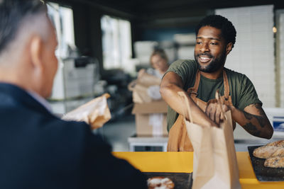 Male baker packing buns in paper bag for customer at bakery