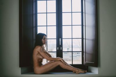 Side view of sensuous naked female model sitting on window sill