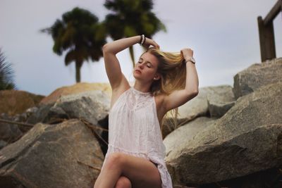 Young woman adjusting hair while sitting on rock against sky