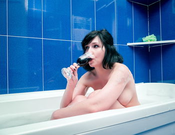 Naked woman drinking wine while sitting in bathtub