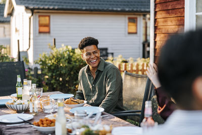 Happy young man sitting on chair near dining table in back yard at dinner party