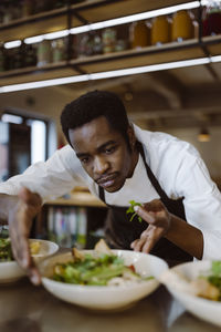 Focused male chef garnishing salad bowl while bending in commercial kitchen