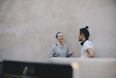 Smiling computer programmer holding smart phone while discussing with male colleague against wall in office