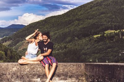 Woman kissing man while sitting on retaining wall