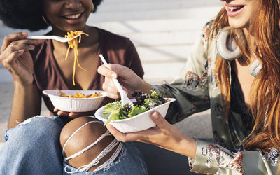 Smiling young female friends eating salad and spaghetti on sunny day