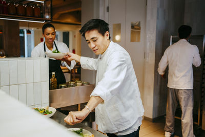 Male chef passing salad bowl to female coworker while finishing salad at commercial kitchen