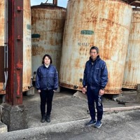 能登半島地震で傾いた焼酎づくりのタンクの前に立つ、日本醗酵化成社長の藤野浩史さん（右）と取締役の裕子さん＝石川県珠洲市で2024年3月（同社提供）