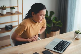 A woman in front of a desk holds her stomach in discomfort. 