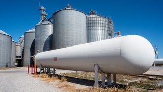 A Large, long white anhydrous ammonia tank with corn grain bins in background at a grain elevator company.