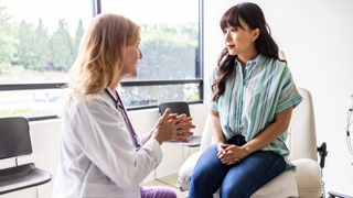 Female patient is shown sat on a white chair at the doctor's office. She has her hands crossed on top of her lap and is looking at a female doctor who appears to be explaining something to her. 