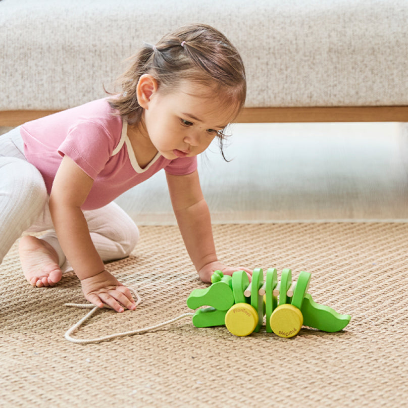 A small child playing with a bright green wooden toy with bright yellow wheels that is shaped like a crocodile.