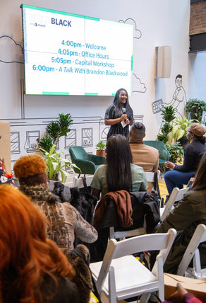 A photo of the Build Black program lead, holding a mic infront of an audience of seated guests at a Build Black event. There's a large monitor behind the program lead indicating the schedule for the event.