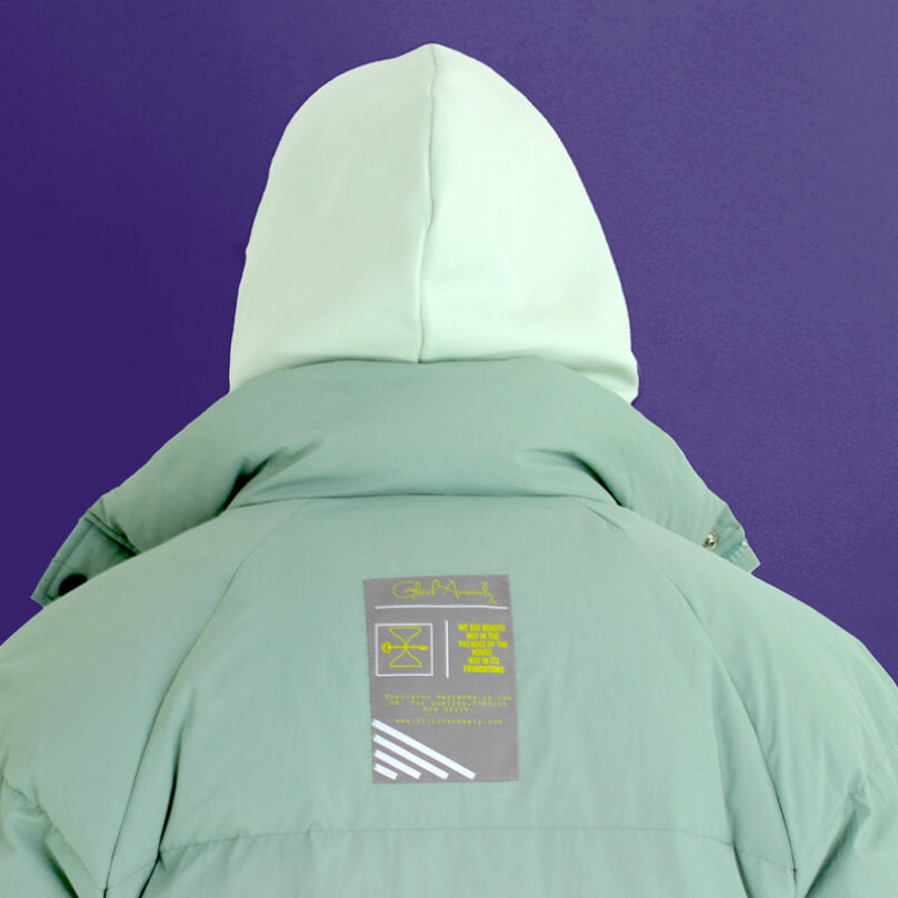 Back of a teal colored winter coat with a lighter hood, featuring a label from Glitch Anomaly.