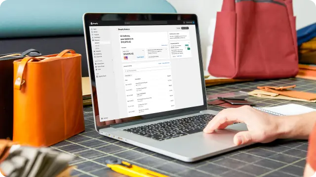 A laptop displaying a Shopify Balance dashboard is open on a dark desk. Colored bags sit on the desk on both sides of the laptop.