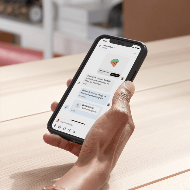 La propietaria de un negocio está sentada frente a un escritorio en su oficina y utiliza Shopify Inbox para chatear con un cliente en su celular. Le comparte un código de descuento en el chat para concretar la venta.