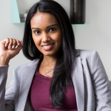 The image features a smiling woman sitting in a chair, wearing a gray suit and a red blouse. She is posing for a photo, and her hands are raised in a confident pose. The woman appears to be a professional, and her attire suggests a formal setting.